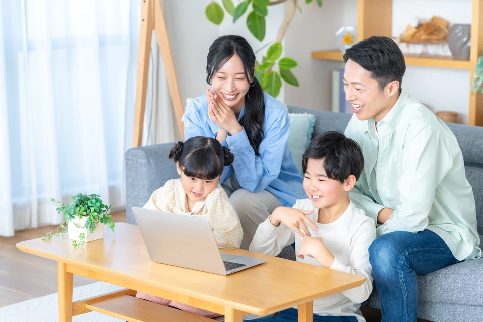 リビングでノートパソコンを囲み、家族4人が笑顔で会話をしている様子